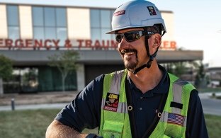 Two people in construction PPE standing outside in front of an emergency and trauma center. 