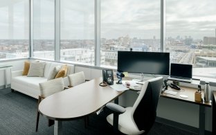 An office with large windows and a desk and monitor set up